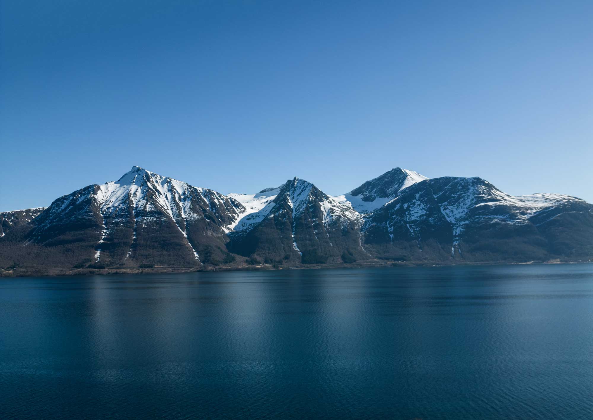 Fjelltopper under blå himmel med vann i forgrunnen som speiler fjellene.