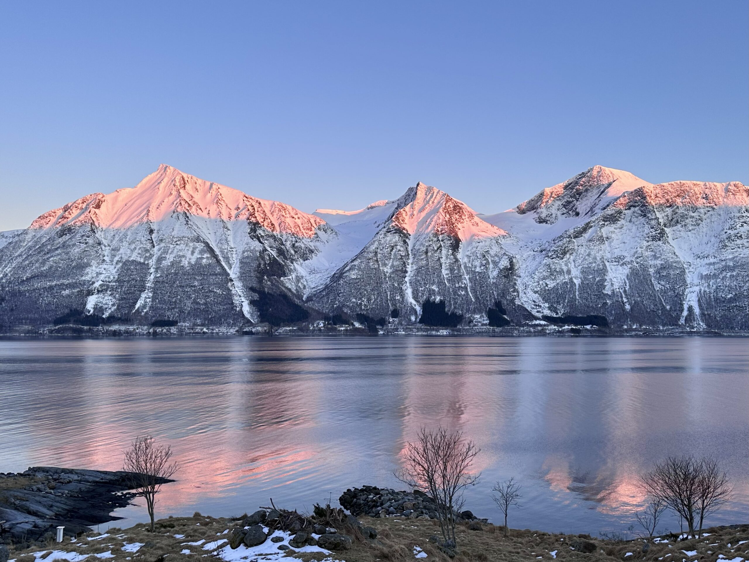 Snødekte fjell som reiser seg over en stille fjord.