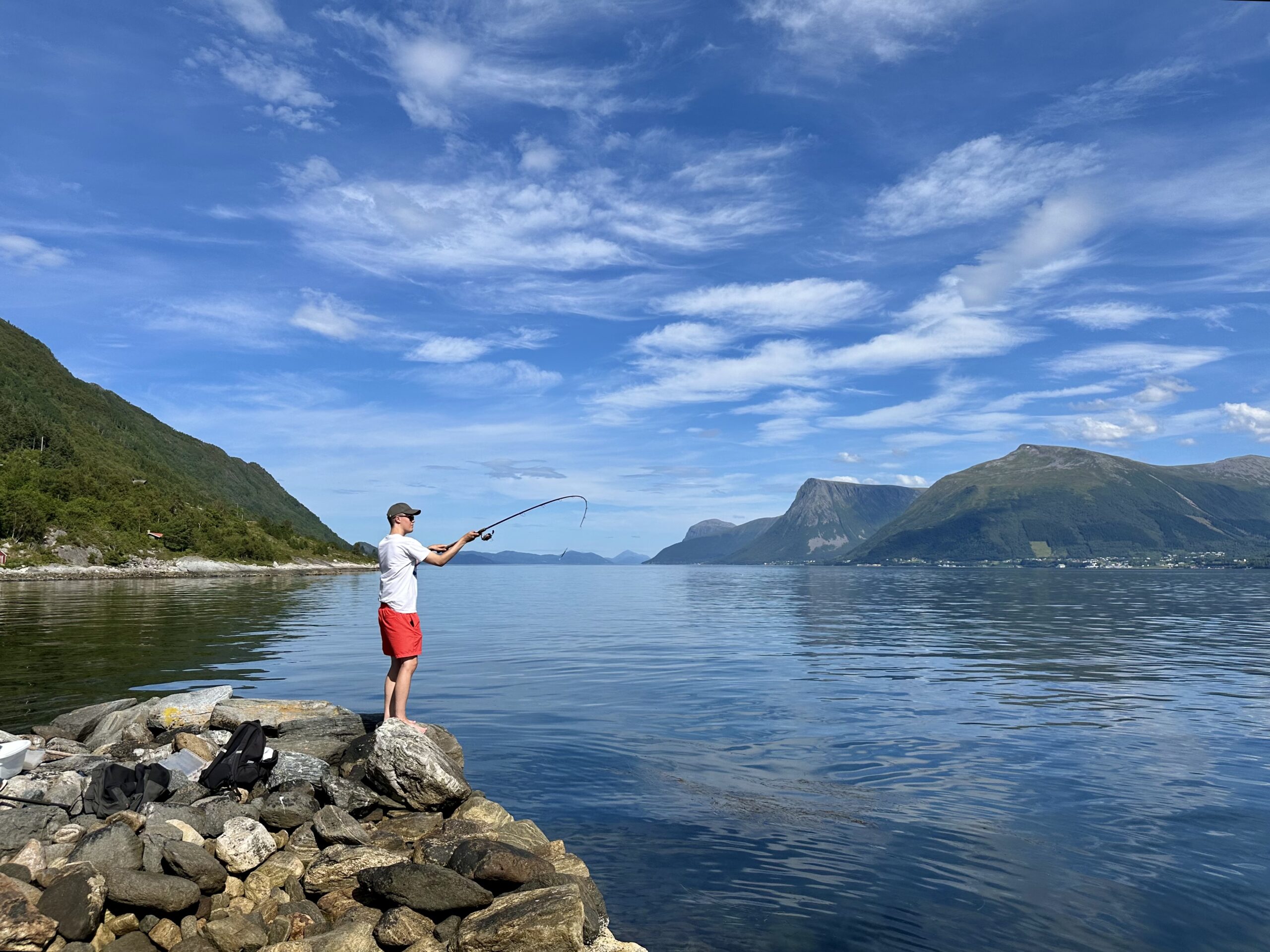 Mann som fisker fra fjordkanten, med vannet og fjell i bakgrunnen.