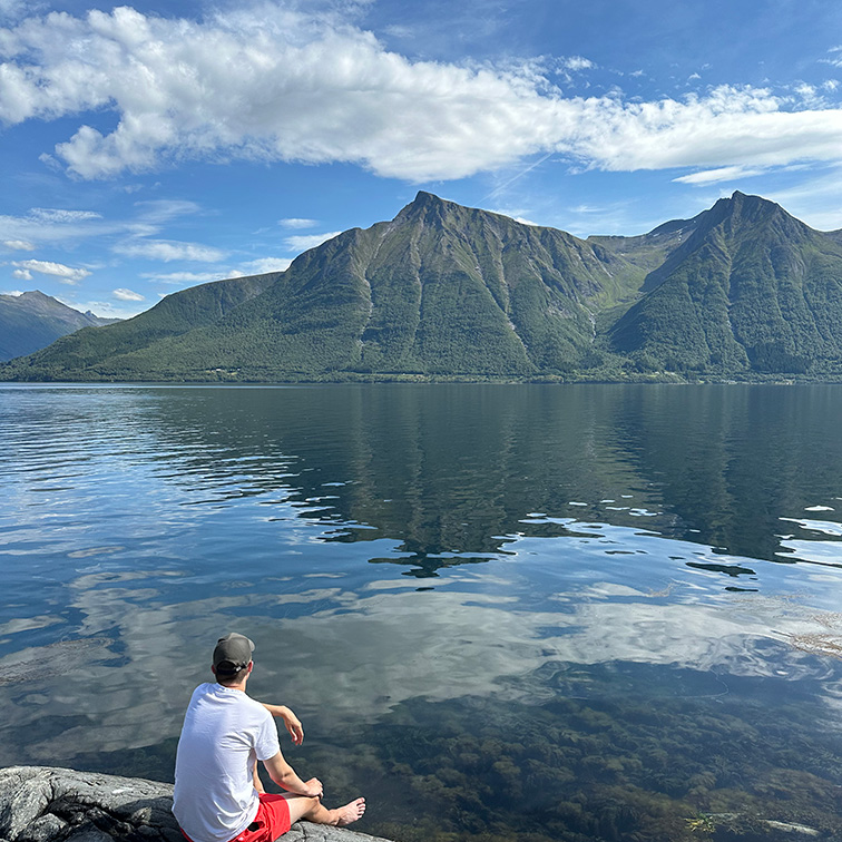 Mann som sitter ved fjordkanten og ser ut over vannet mot fjell på den andre siden, med blå himmel og spredte hvite skyer i bakgrunnen.